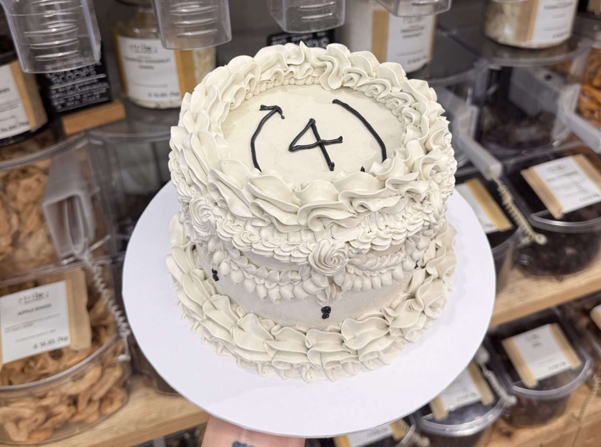 A Lambeth-style celebration cake with elaborate cream-coloured buttercream piping, decorated with the Tribe Zero-Waste recycling arrow logo and the number 4 piped in black icing on top. The cake is held up against a backdrop of Tribe Zero-Waste's loose goods dispensers and shelving, filled with nuts, grains and dried fruit.